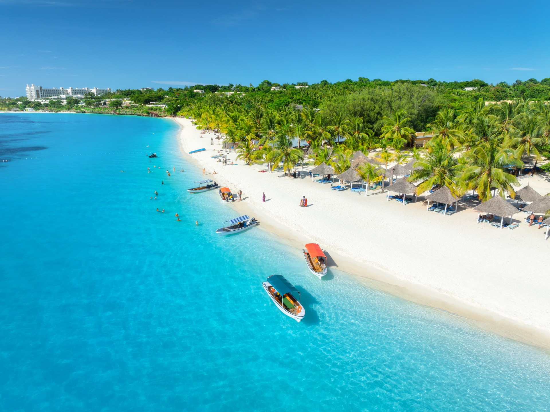 Aerial view of the boats on tropical sea coast with white sandy beach at sunset. Summer holiday in Kendwa, Zanzibar. Landscape with boat, yacht, clear blue water, green palm trees. Top drone view